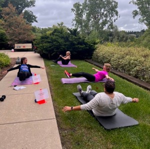 Instructor with 3 people outdoors on grass in a yoga post with arms and legs raised while on yoga mats