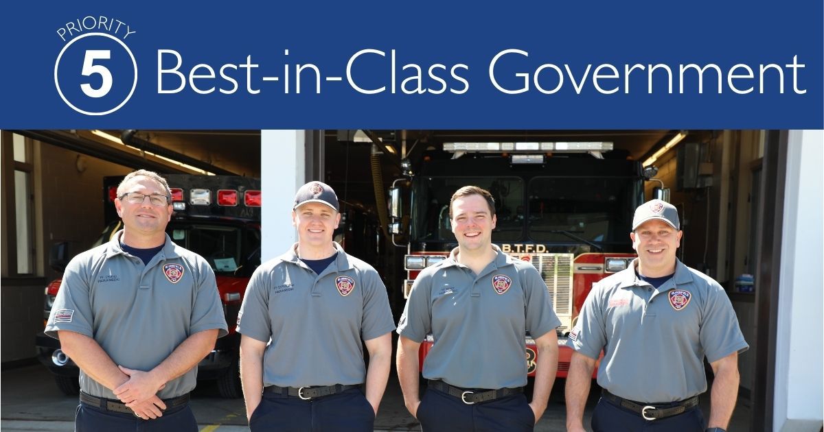 Four firefighters standing in front of the fire station with fire trucks parked inside. Title of Priority 5: Best-in-Class Government