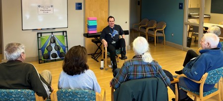 Group of seniors in chair doing exercises with their legs that match instructor. Room is equipped with colorful blocks, a toss game, and a sign that reads "Welcome to the Friendship Club"