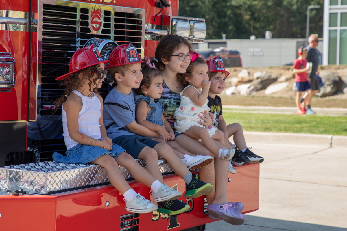 Six kids wearing plastic fire hats sitting on the bumper of a red fire truck
