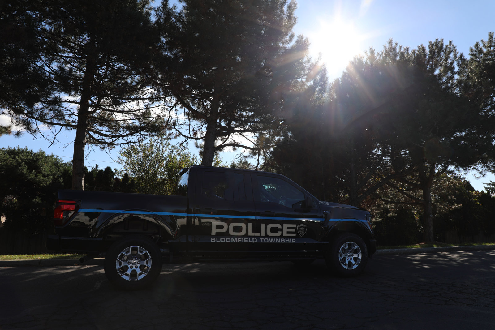 Dark pickup truck with Police Bloomfield Township written on side parked next to a row of tall pine trees and the sun glaring brightly in the sky.