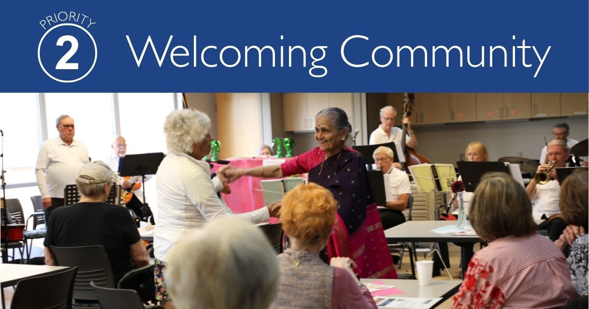 Two ladies holding hands while dancing and smiling with onlookers and a band playing in the background inside the Senior Center. Title Priority 2: Welcoming Community